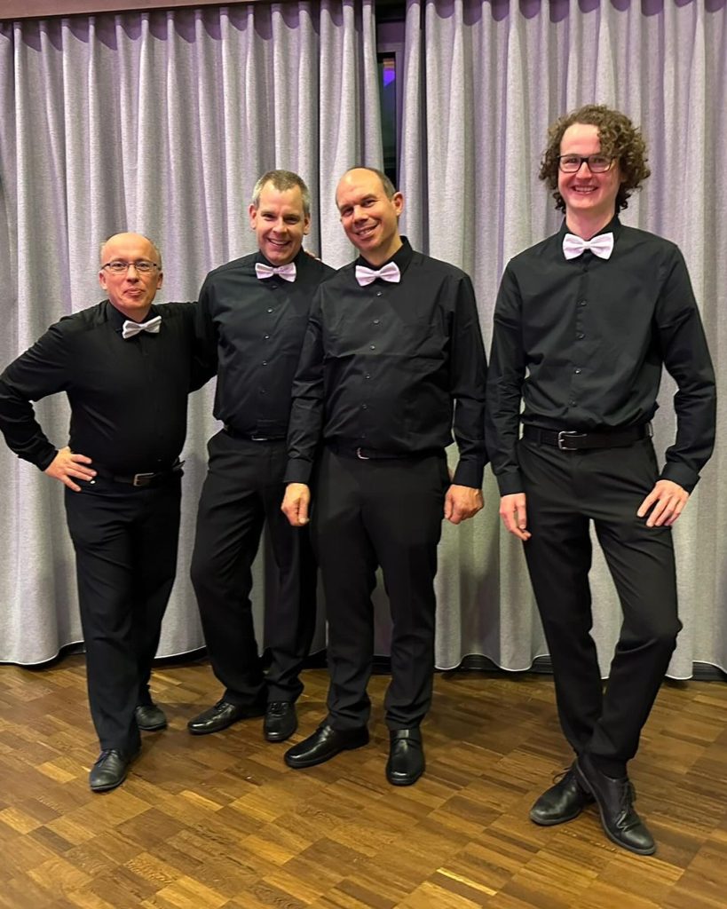 Vier Musiker in schwarzen Hemden mit Fliege beim Konzert 2023